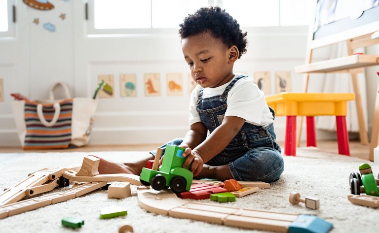 Infant playing with wooden train toys on a carpet in a bright classroom.