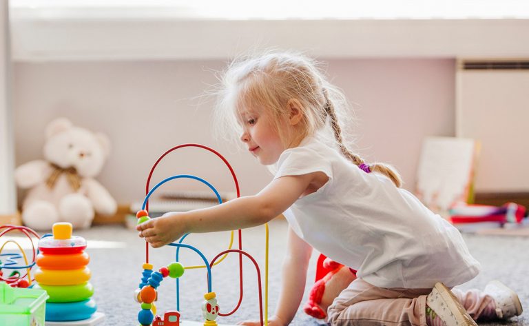 Toddler playing with colorful bead maze toy on the floor in a daycare classroom.